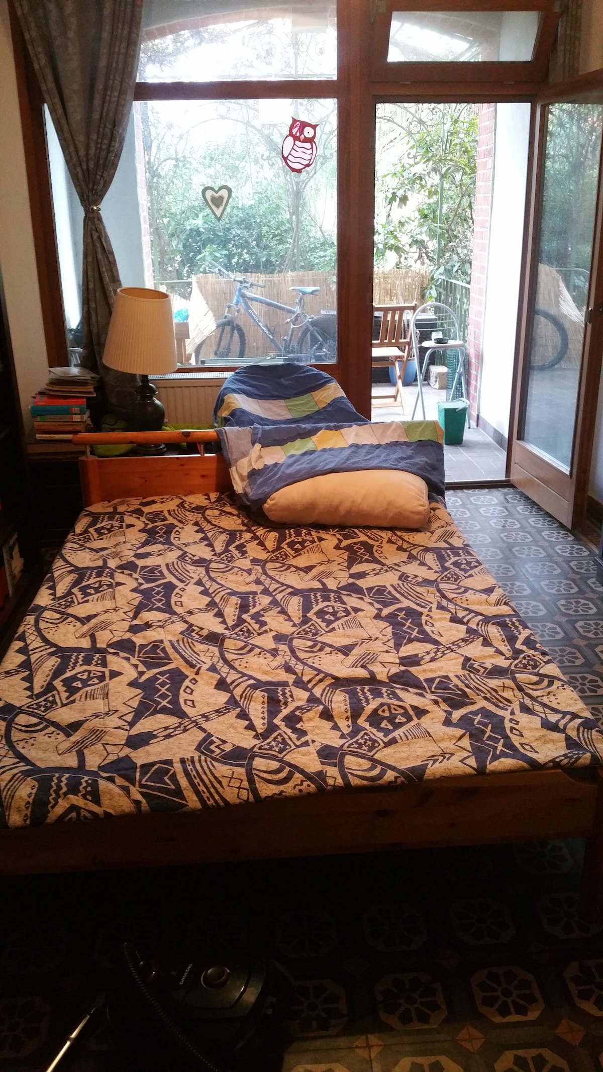 A spacious bed with a patterned blanket is positioned in a cozy room. Natural light filters through glass doors leading to an outdoor area. A small lamp sits on a wooden bedside table, while books are neatly arranged on a nearby shelf.
