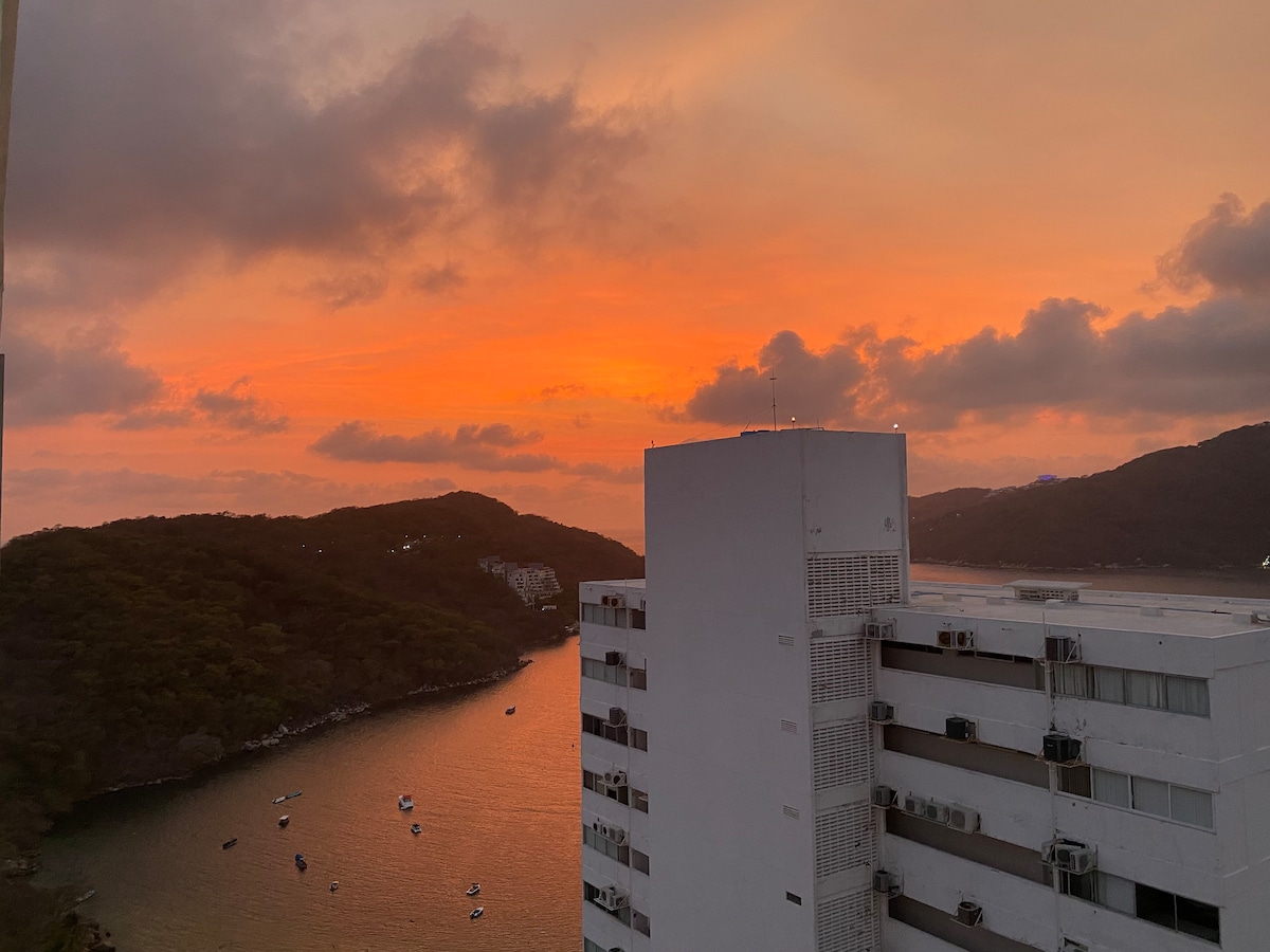A stunning sunset paints the sky in shades of orange and pink over a tranquil bay. Silhouettes of distant hills and buildings are visible, alongside calm waters reflecting the vibrant hues of evening. A peaceful ambiance is created by the serene landscape.