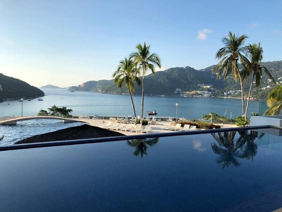 An expansive view of a tranquil bay is captured, with lush green hills framing the water. Palm trees are seen swaying gently, while a serene infinity pool reflects the sky. Sun loungers are arranged around the pool, inviting relaxation by the serene surroundings.