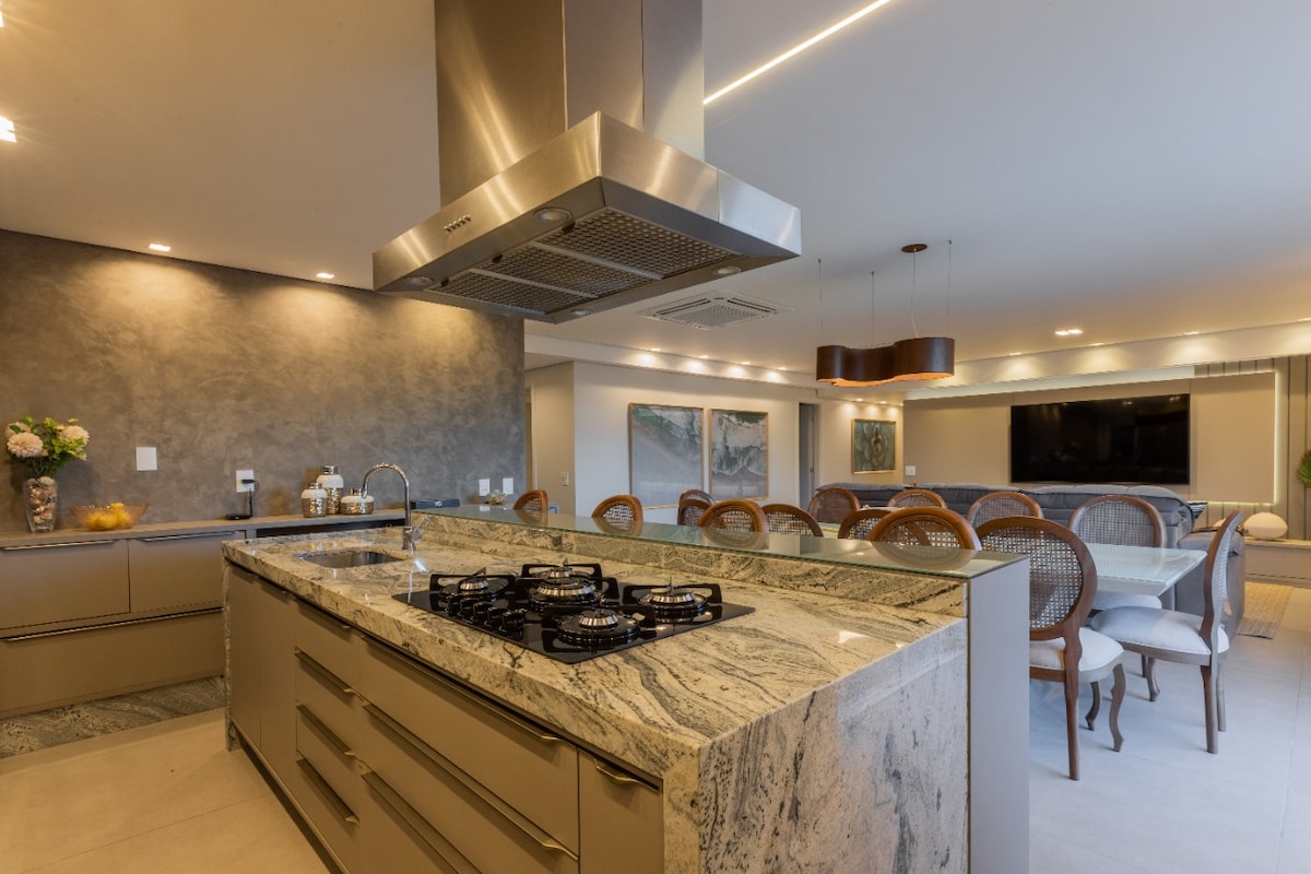 The kitchen features a spacious island with a marble countertop, equipped with a gas cooktop. Soft lighting highlights modern cabinetry. In the background, a dining area is visible with multiple dining chairs and a large table, creating an inviting space for meals.