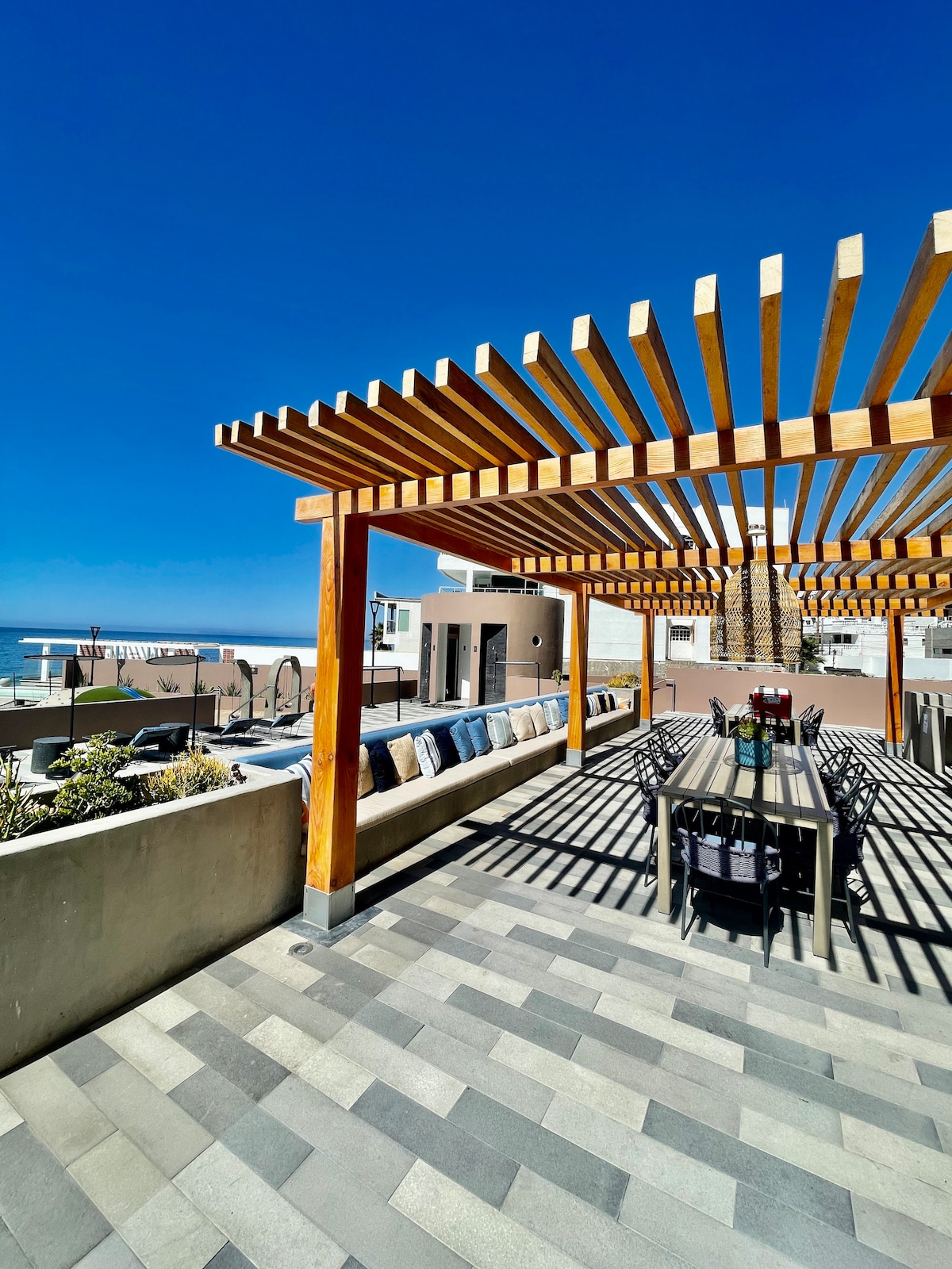 A spacious outdoor terrace is shaded by a wooden pergola, with comfortable seating arranged along the wall. A long dining table is positioned centrally, offering a view of the ocean and the clear blue sky above. Paved flooring creates a modern aesthetic.