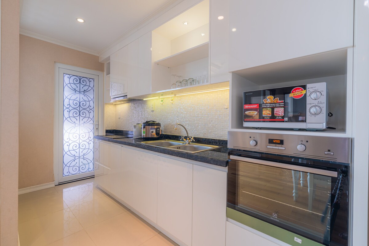 A modern kitchen is equipped with sleek white cabinetry and a black countertop. Stainless steel appliances, including a microwave and an oven, are neatly arranged. Soft lighting illuminates the space, and a decorative glass door adds an elegant touch.
