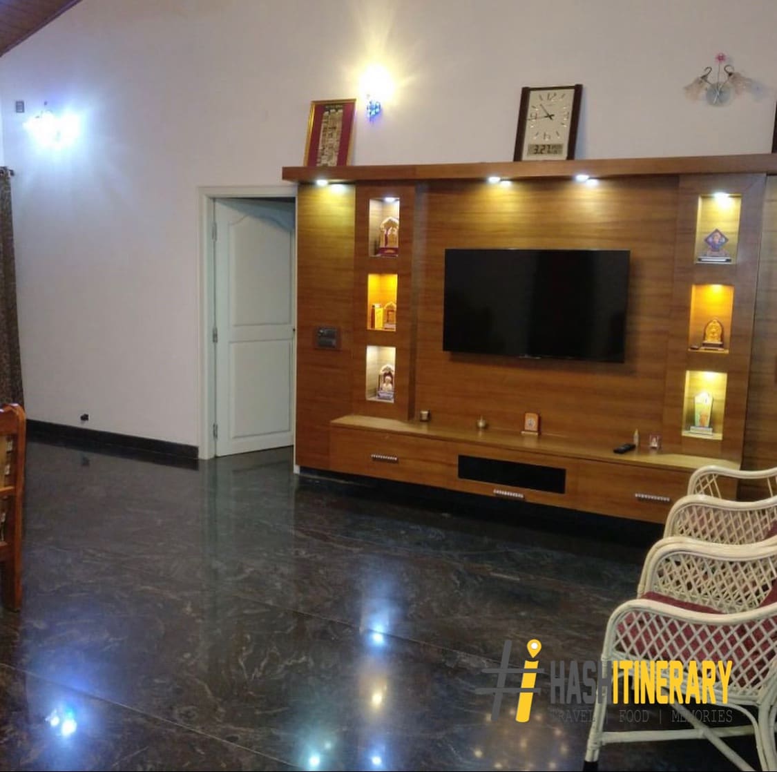 A spacious living area features polished black marble flooring and a wooden entertainment unit with a wall-mounted television. Decorative items are displayed on the unit, and soft lighting is provided by wall sconces. An adjacent doorway leads to another room.