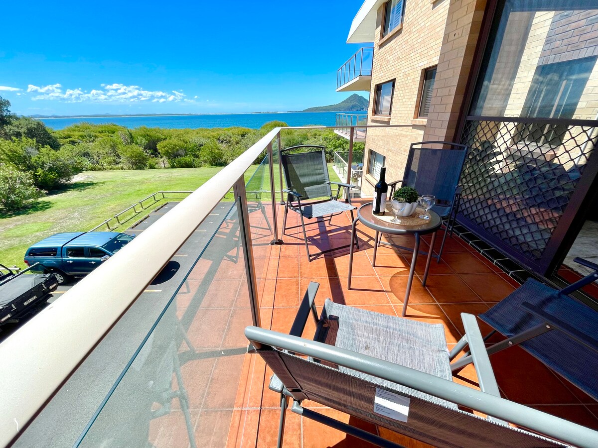 A private balcony is shown with two chairs and a small table, featuring scenic views of the water and greenery. The space is identified by terracotta tiles and a glass railing, allowing for unobstructed views of the surroundings.