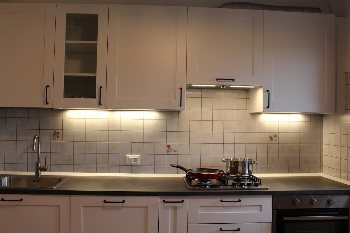 A modern kitchen is presented with sleek white cabinets and a clean gray countertop. Under-cabinet lighting illuminates the space, highlighting a stove and oven set. A stainless steel sink is positioned beside the stove, while a pot sits on the burner, indicating a functional cooking space.
