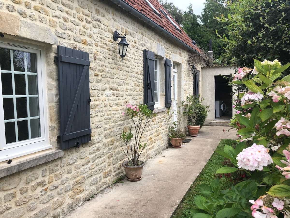 A charming stone exterior is highlighted by gray shutters and a lantern. Flower pots with greenery flank a pathway leading to an entrance. Lush bushes with pink flowers and greenery enhance the serene ambiance of the surrounding landscape.