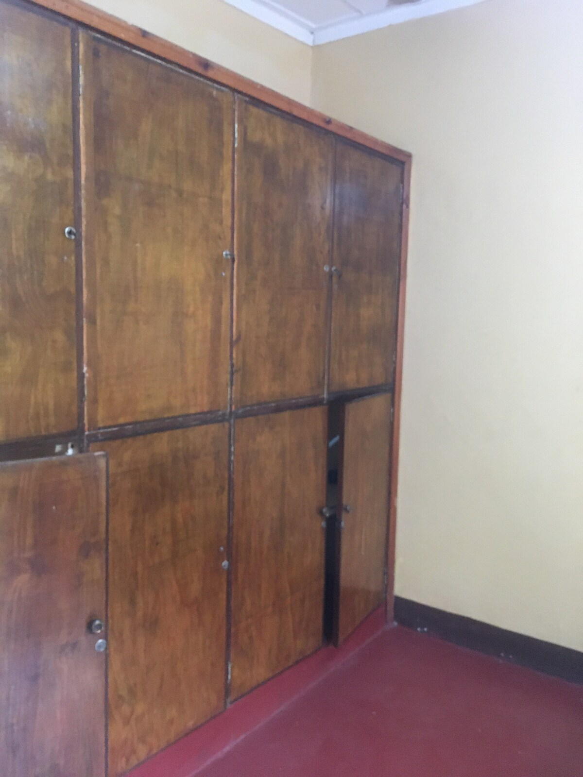 A wooden wardrobe features several doors, showcasing a warm, natural finish. One door is opened, revealing additional storage space inside. The surrounding wall is painted in a soft yellow hue, complemented by a red floor, creating a cozy atmosphere.