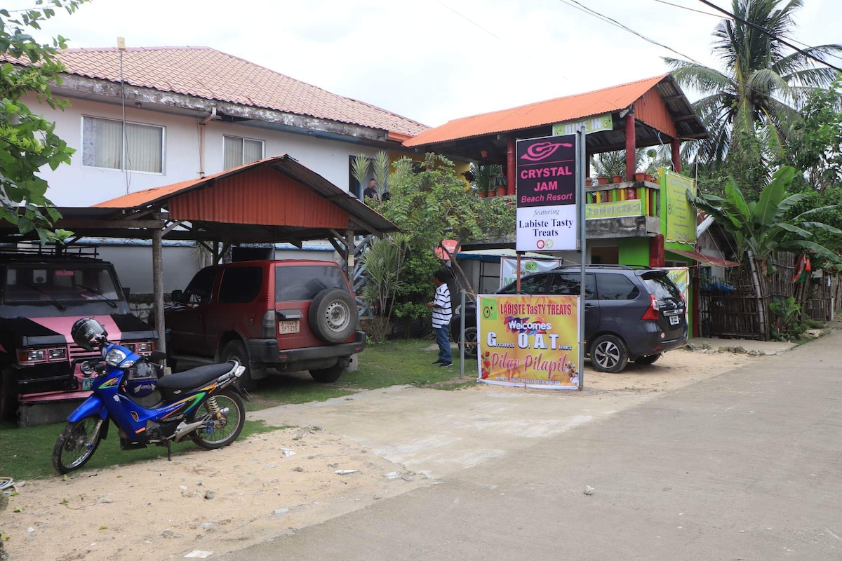 A beachfront resort is shown with a bright sign reading 'Crystal Jam.' Several vehicles are parked beneath the shaded area, including a motorcycle and cars. Lush greenery and palm trees surround the property, creating a relaxed outdoor space.