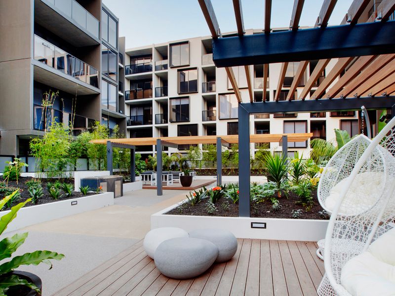 A communal terrace area is depicted, featuring landscaped gardens with various plants, shaded seating spaces, and unique furniture. Multiple levels of greenery enhance the space, while an elegant structure of wooden beams provides a modern aesthetic. The inviting layout encourages relaxation and socializing.