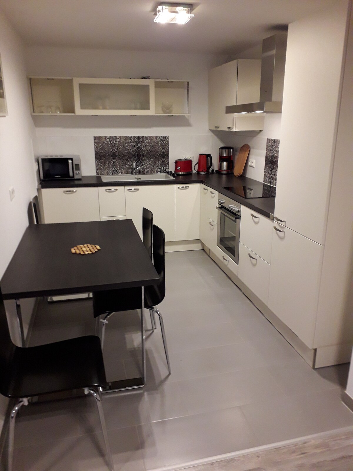 A functional kitchen is equipped with modern appliances, including a microwave and a coffee maker. A dining table with chairs is positioned next to the cabinetry. Neutral-colored cabinets contrast with the dark countertops, while a decorative item is placed on the table.