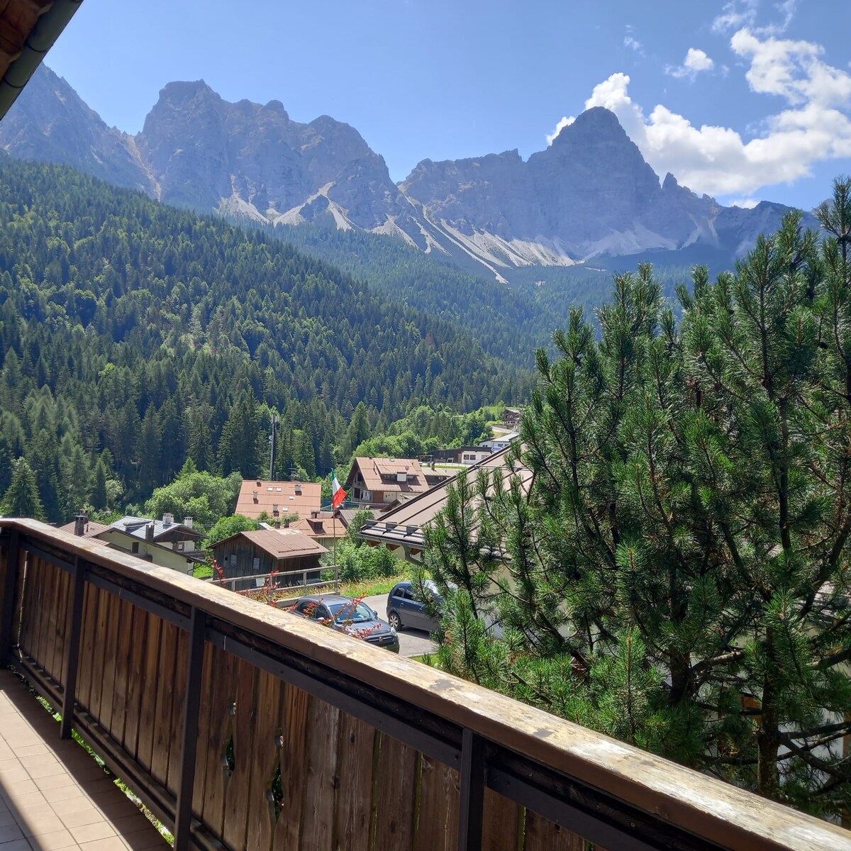 A view from the balcony showcases a mountainous landscape with towering peaks and lush forests. The foreground contains a wooden railing with hanging plants. Below, traditional chalets are visible, with a flag gently fluttering in the breeze amongst the greenery.