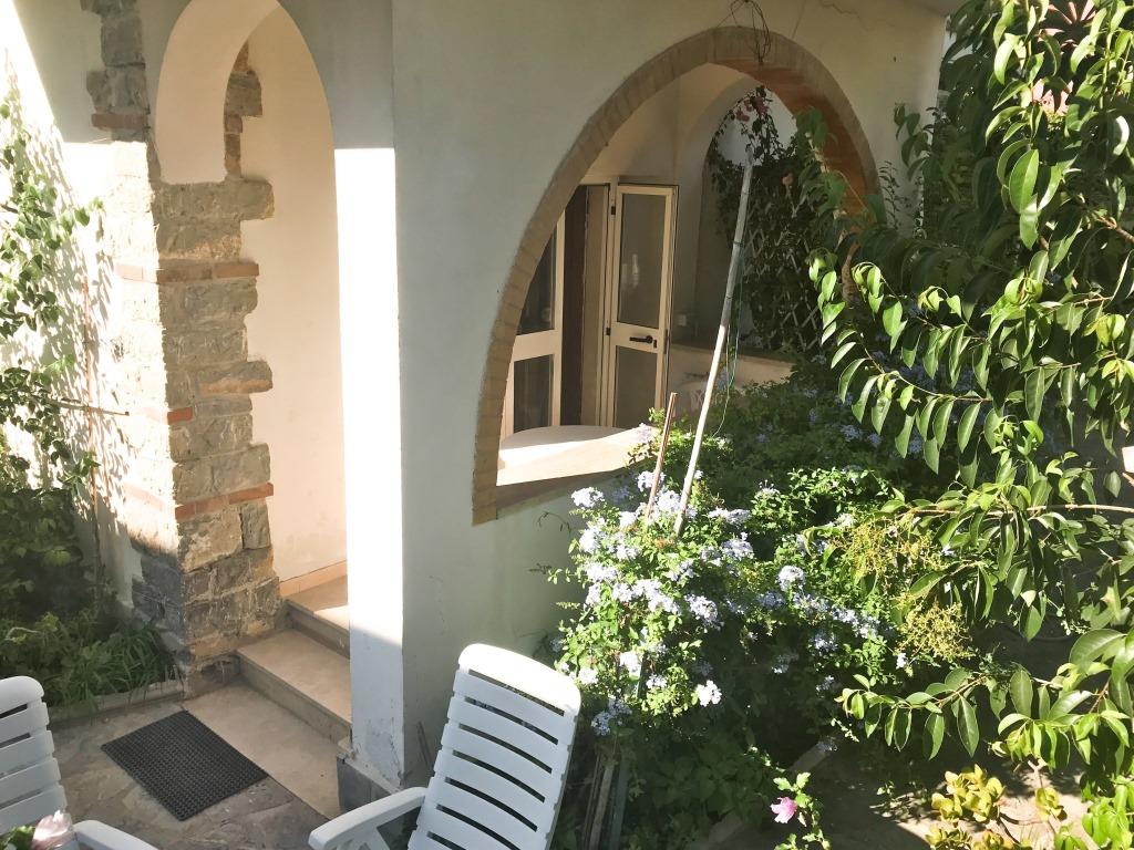 An inviting outdoor area is framed by lush greenery and flowering plants. A pathway leads to a welcoming entrance, while white lounge chairs are arranged on a tiled patio, providing a comfortable space to relax in nature.