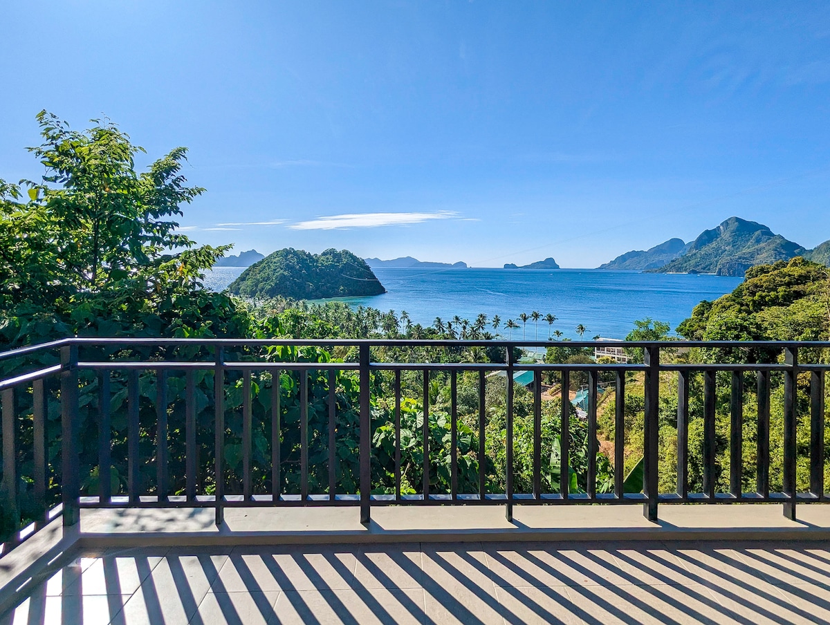 A spacious balcony showcases expansive views of Bacuit Bay and surrounding islands. Lush greenery frames the scene, complementing the calm blue waters. The clear sky enhances the serenity of the landscape, creating a tranquil atmosphere for relaxation.