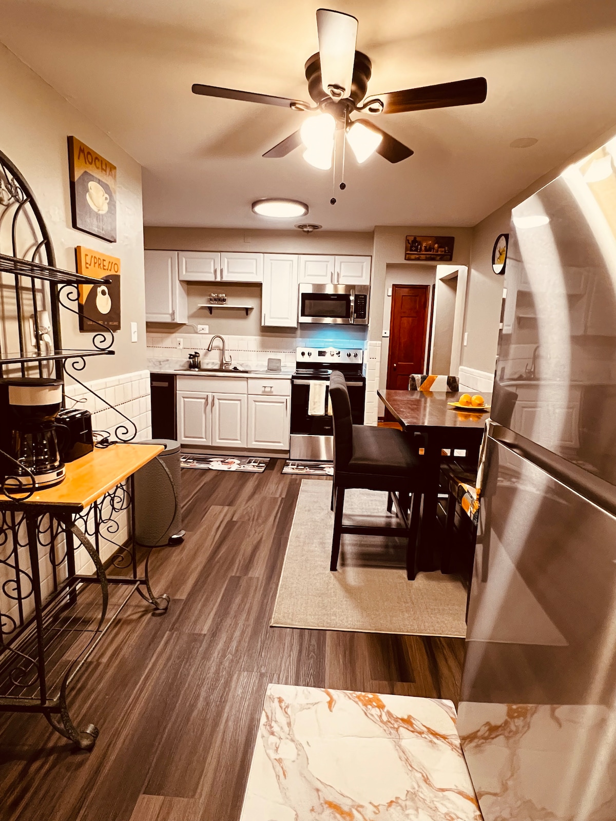 The modern kitchen features light cabinetry and stainless steel appliances, including a refrigerator and oven. A small dining table with two black chairs occupies the space. A ceiling fan provides ventilation, while warm lighting enhances the inviting atmosphere.