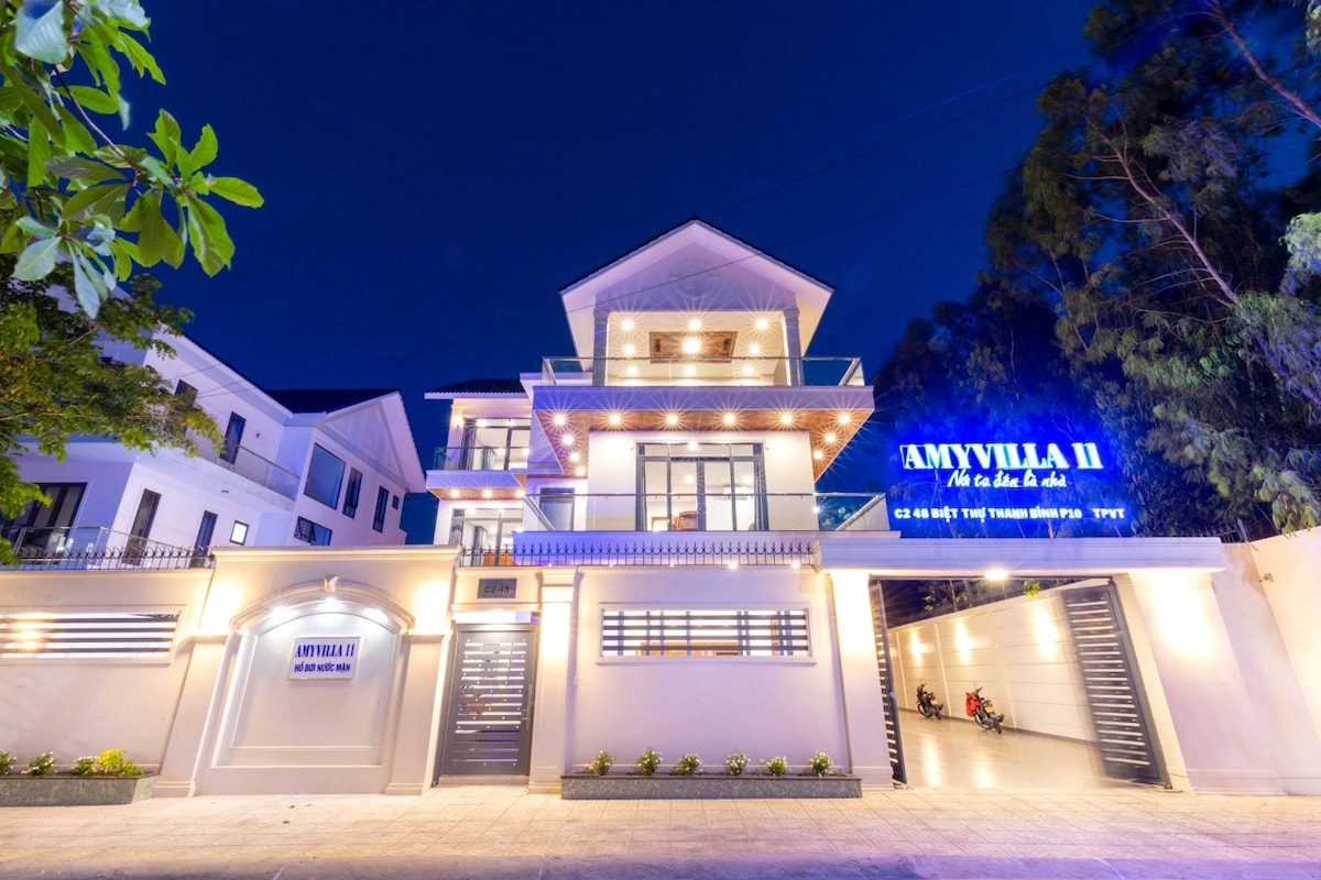 The villa's exterior is illuminated by soft lighting in the evening, highlighting the modern architectural design. A welcoming entrance is complemented by neatly arranged greenery and a spacious driveway, providing access to the property.