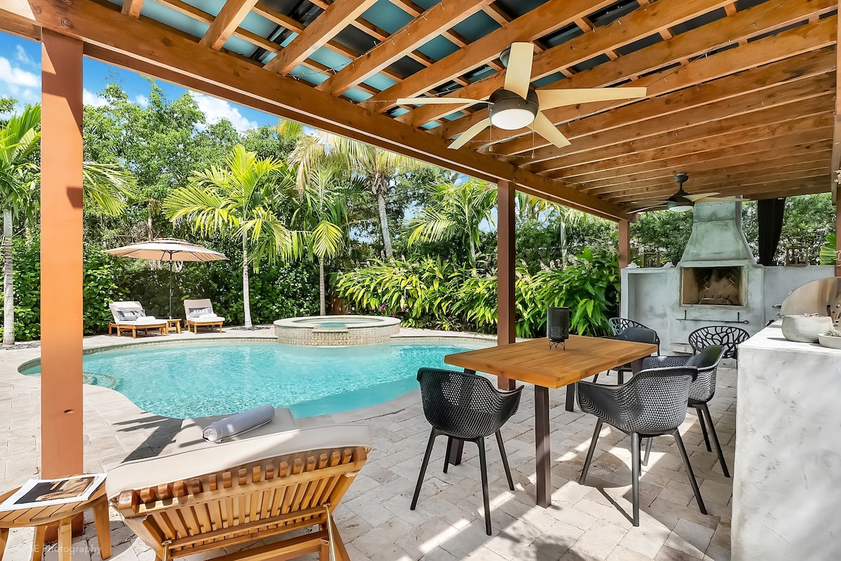 An outdoor dining area is set beneath a wooden pergola, featuring a table surrounded by black chairs. A refreshing heated pool is seen alongside a spa and lounge chairs, with lush tropical foliage providing privacy and shade.