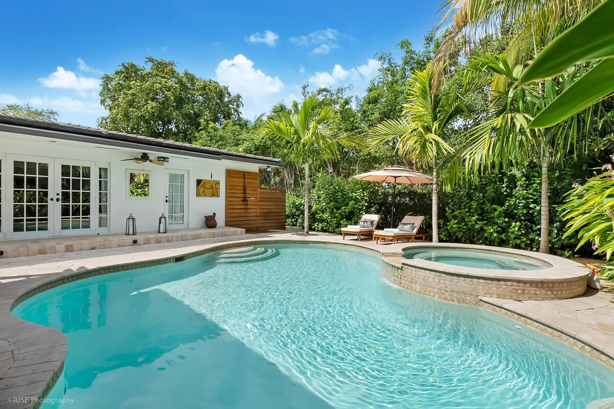 A heated private pool encircled by lush tropical foliage provides a serene outdoor retreat. The pool area features a separate spa, lounge chairs, and an umbrella for shade, along with a wall that enhances privacy and tranquility.