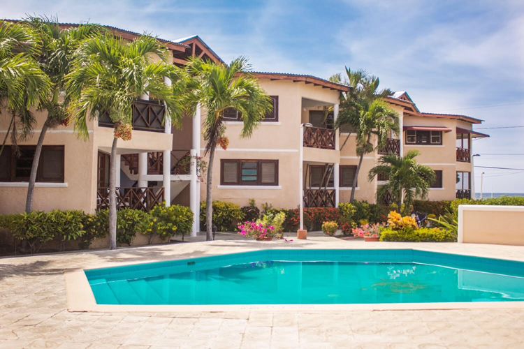 A pool area is framed by lush palm trees and vibrant landscaping, featuring a clear blue swimming pool. The three-story building showcases a light-colored facade with balconies, harmonizing with the tranquil outdoor space.