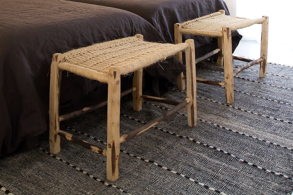 Two woven stools are positioned beside a bed with a dark brown coverlet. The stools feature a natural wood finish, highlighting a rustic design. A textured, patterned area rug complements the scene beneath the furniture, enhancing the room's aesthetic.