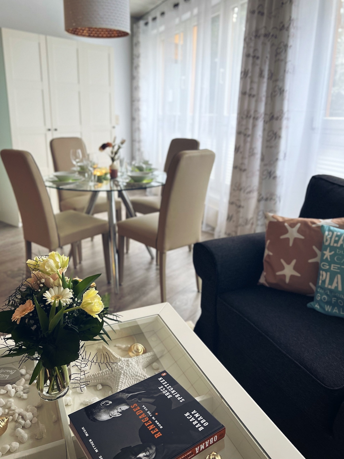 A dining area features a round glass table surrounded by four contemporary chairs, set for a meal. A cozy seating area includes a dark sofa adorned with decorative pillows. A floral arrangement and a few books are displayed on a coffee table, adding charm to the space.