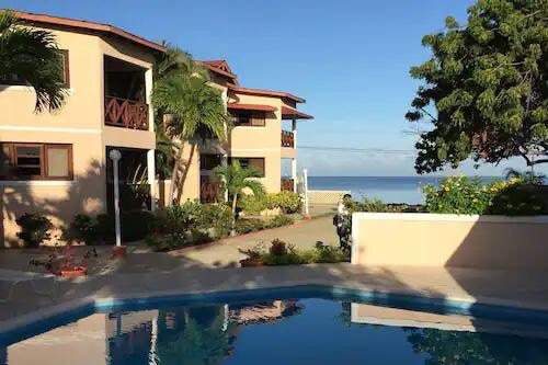 A two-story building is set against a clear blue sky, nestled among palm trees and vibrant greenery. The outdoor space features a serene pool reflecting the surroundings, while the ocean is visible in the background, enhancing the inviting environment.