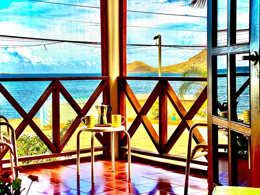 A sunlit balcony is captured, showcasing a scenic view of the ocean and distant hills. A small table is set with two yellow cups and a coffee pot. Wooden accents frame large windows, inviting natural light into the space.