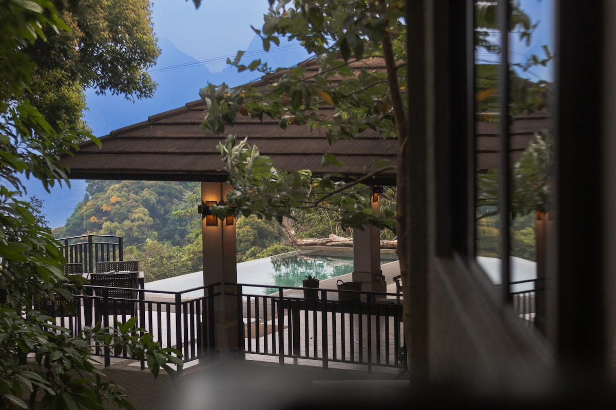 The exterior lounge area is framed by lush greenery, providing views of a tranquil infinity pool. The roof features warm lighting, creating a cozy ambiance. Surrounding seating arrangements invite relaxation while being enveloped by the natural beauty of the landscape.