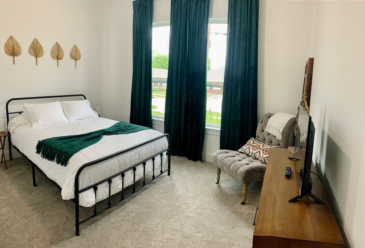 A bright room features a comfortable bed with an inviting white duvet and a textured green throw. Large windows allow natural light to fill the space, complemented by dark curtains. A plush chair and a wooden TV stand offer additional seating and entertainment options.