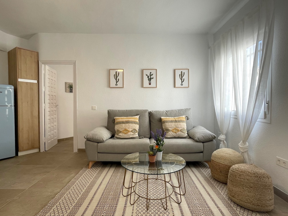 A living area features a light gray sofa with accent pillows, positioned before a glass coffee table. A small potted plant rests on the table. Two natural woven poufs are placed on a patterned area rug, while sheer white curtains frame a window allowing natural light.