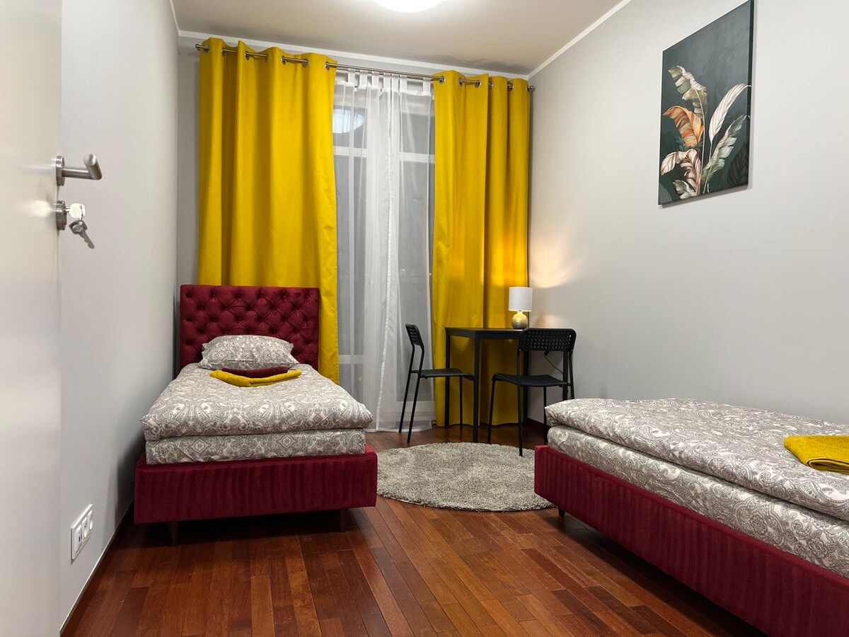 A bright room features two single beds with patterned bedding and plush accents. Large yellow curtains frame a window, complementing the warm wood flooring. A small round table with two black chairs is positioned near the window, while a simple artwork adorns the wall.