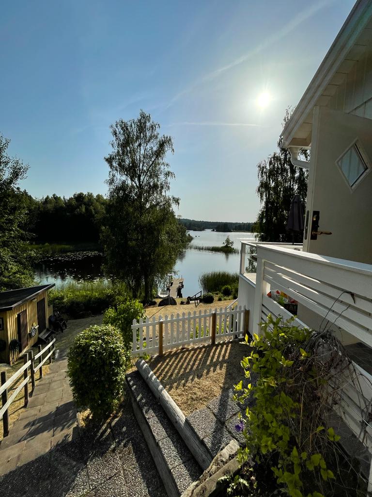 A serene lakeside view is presented, featuring gently sloping steps leading down to the water. Sunlight reflects on the lake's surface, framed by trees and greenery. A wooden dock is visible at the water's edge, enhancing the tranquil setting.