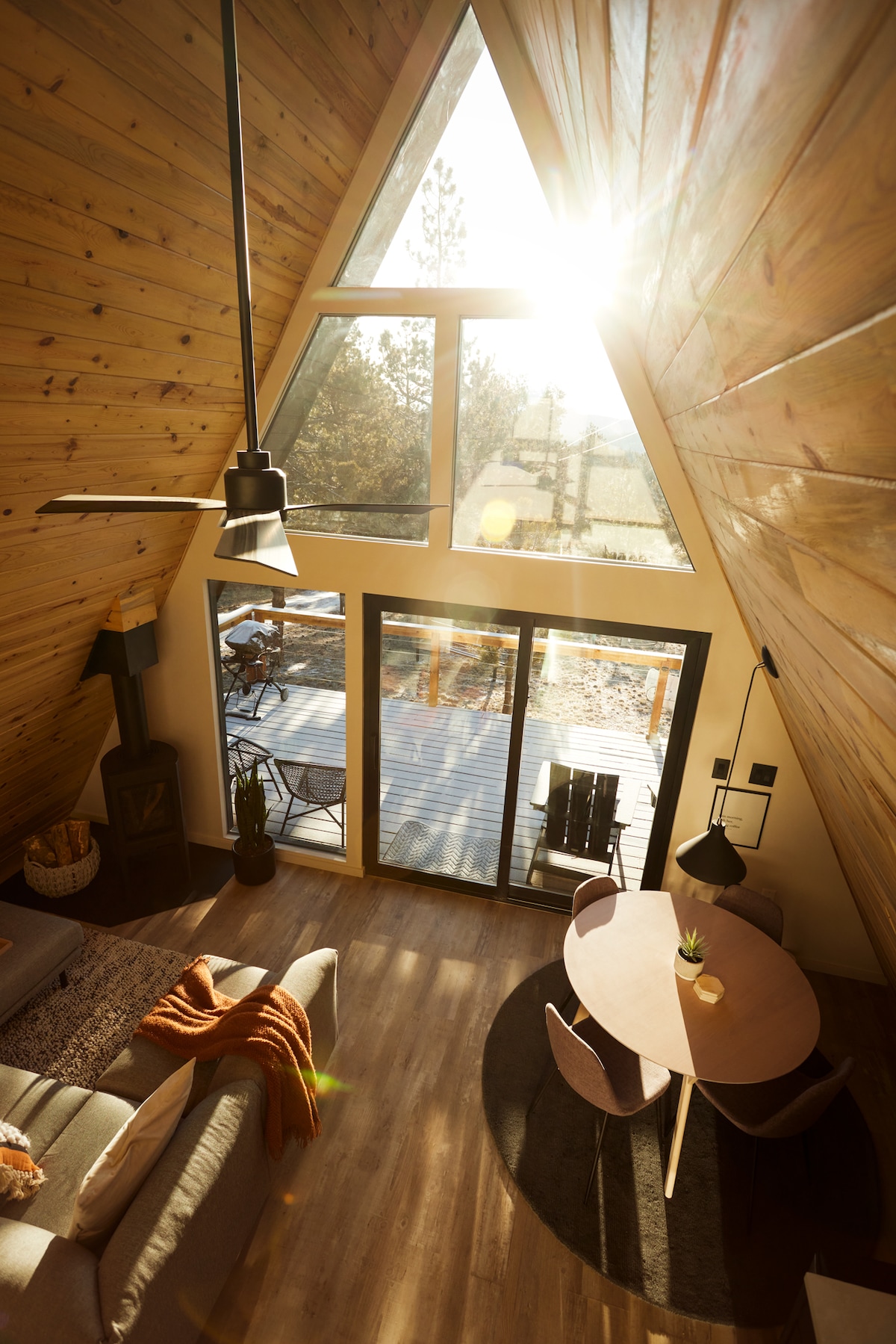 Natural light floods the open living area of the A-frame, highlighting the wooden walls and the modern furnishings. A comfortable sofa is positioned by a cozy gas fireplace, with a round dining table nearby. Large glass doors offer access to the outdoor deck.