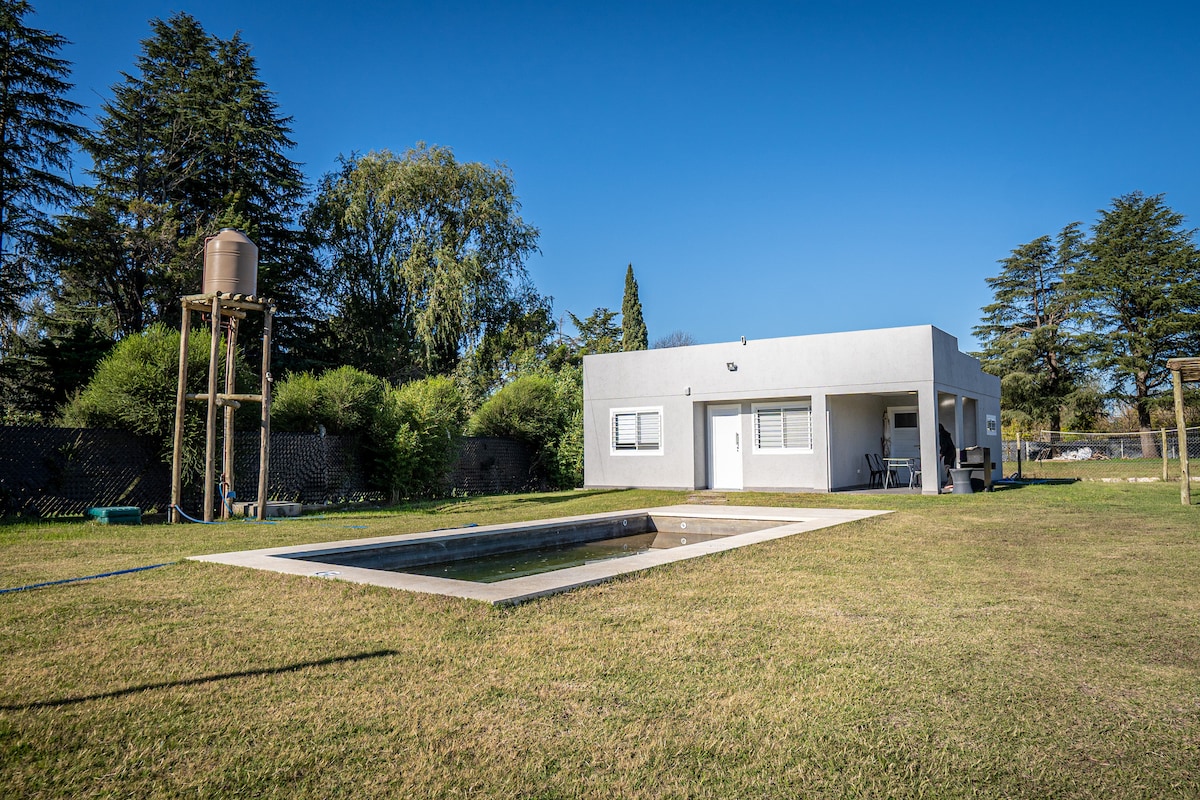 A modern building is situated in a spacious green area, surrounded by trees. A rectangular swimming pool is visible in the foreground, with a utility water tank positioned nearby. Clear blue skies create a serene backdrop for the peaceful setting.