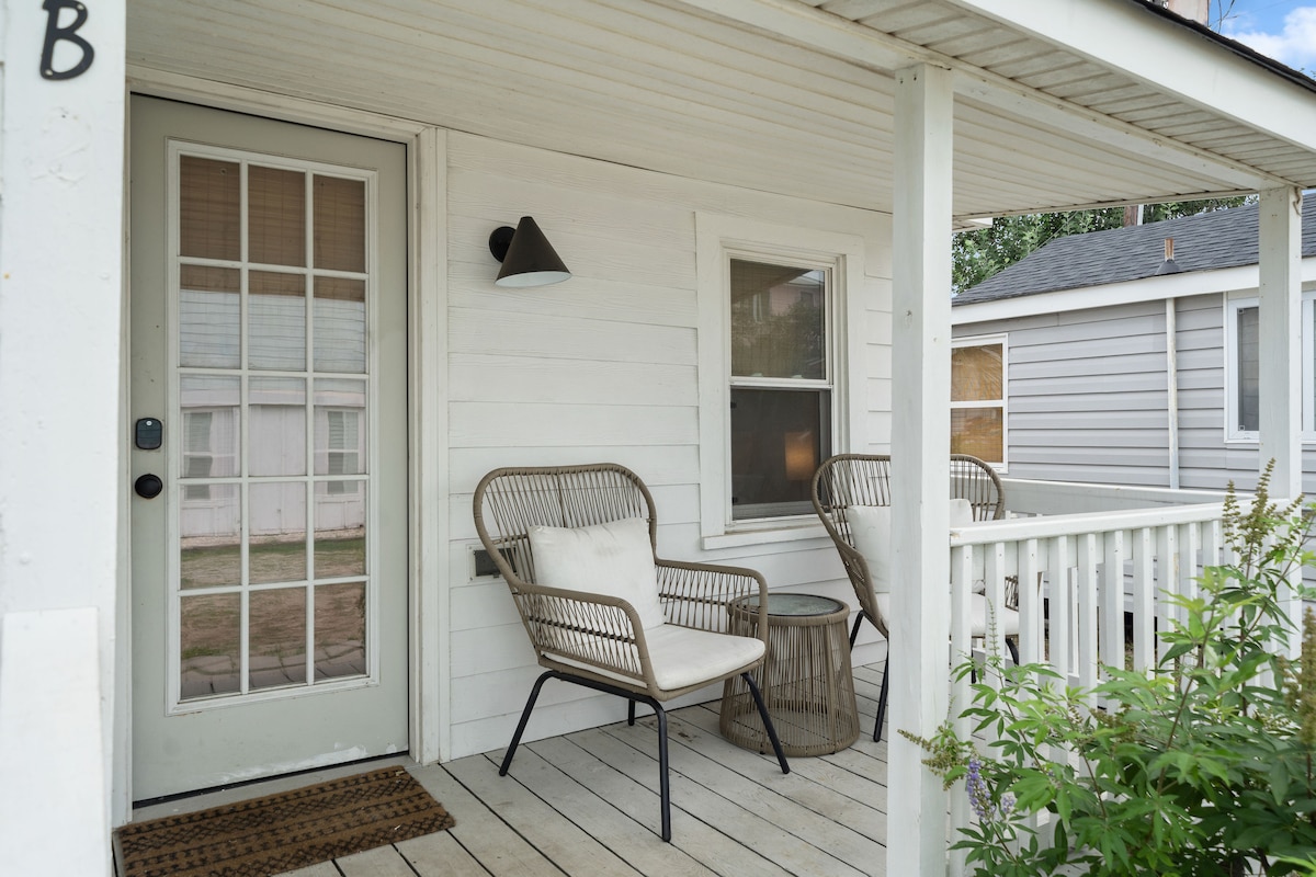 A charming porch area is displayed, featuring two rattan chairs with soft cushions positioned around a small, round table. A wall-mounted light fixture offers understated illumination. The entrance door is framed by glass panes, enhancing the welcoming vibe of this outdoor space.