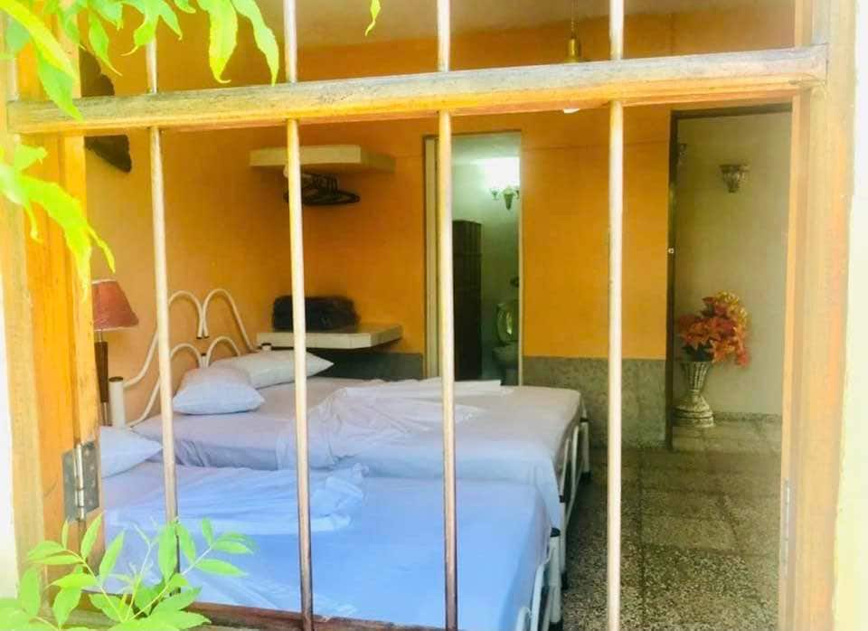 The room is visible through a barred window, showcasing two beds with light blue linens. A compact table with storage is positioned to one side, while a small decorative plant adds a touch of color. A doorway leads to a softly lit area beyond.