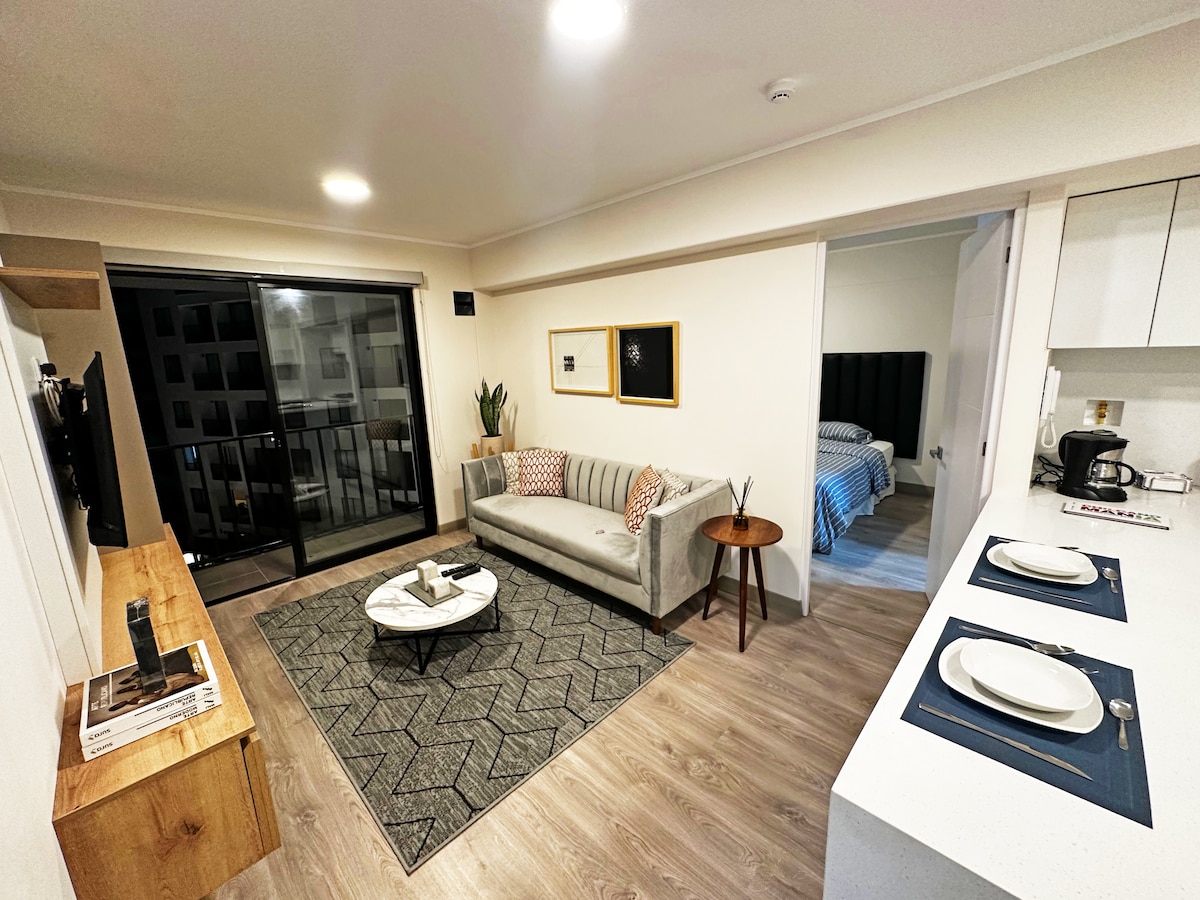 A comfortable living area is observed, featuring a light gray sofa adorned with patterned cushions. A round coffee table sits atop a textured area rug. Bright overhead lights illuminate the space, while the floor-to-ceiling glass door leads to a balcony. The kitchen area is partially visible.
