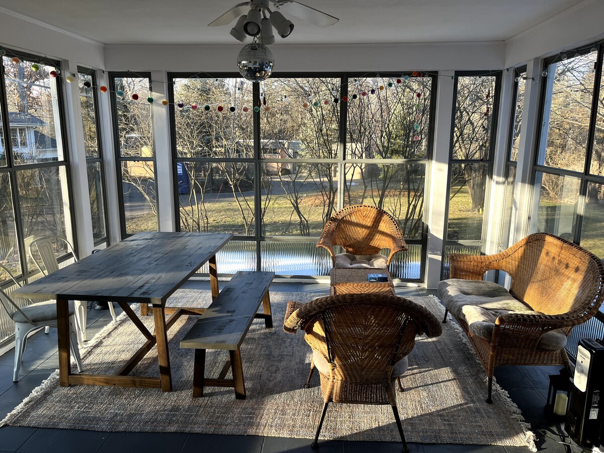 A sun room is filled with natural light, featuring a wooden dining table accompanied by a long bench. Two wicker chairs, adorned with cushions, flank the table. Large windows provide views of the surrounding greenery, while decorative lights hang from the ceiling.