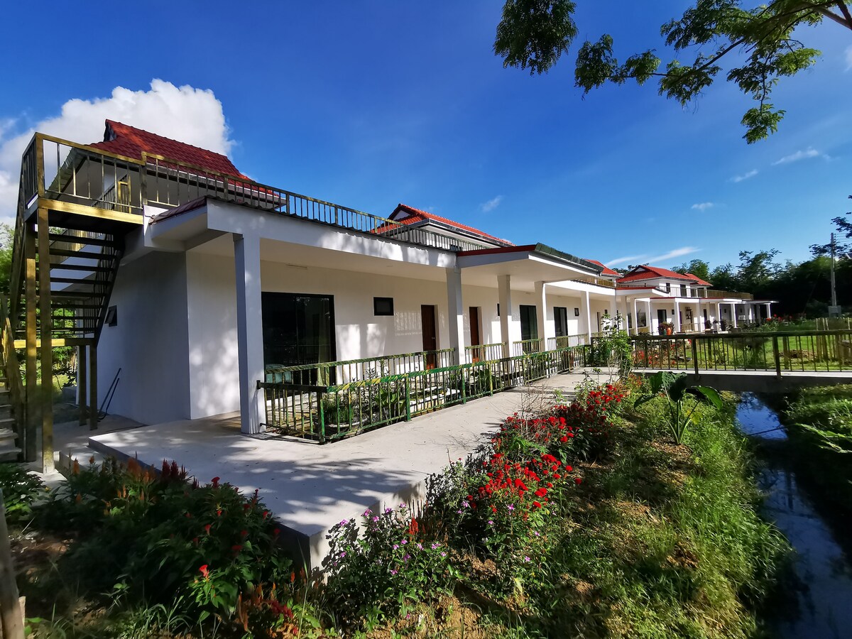 A modern house is showcased with a wide staircase leading to an elevated deck. The exterior features white walls complemented by a red-tiled roof. Lush landscaping with colorful flowers lines the pathways, and a tranquil waterway runs alongside the property.