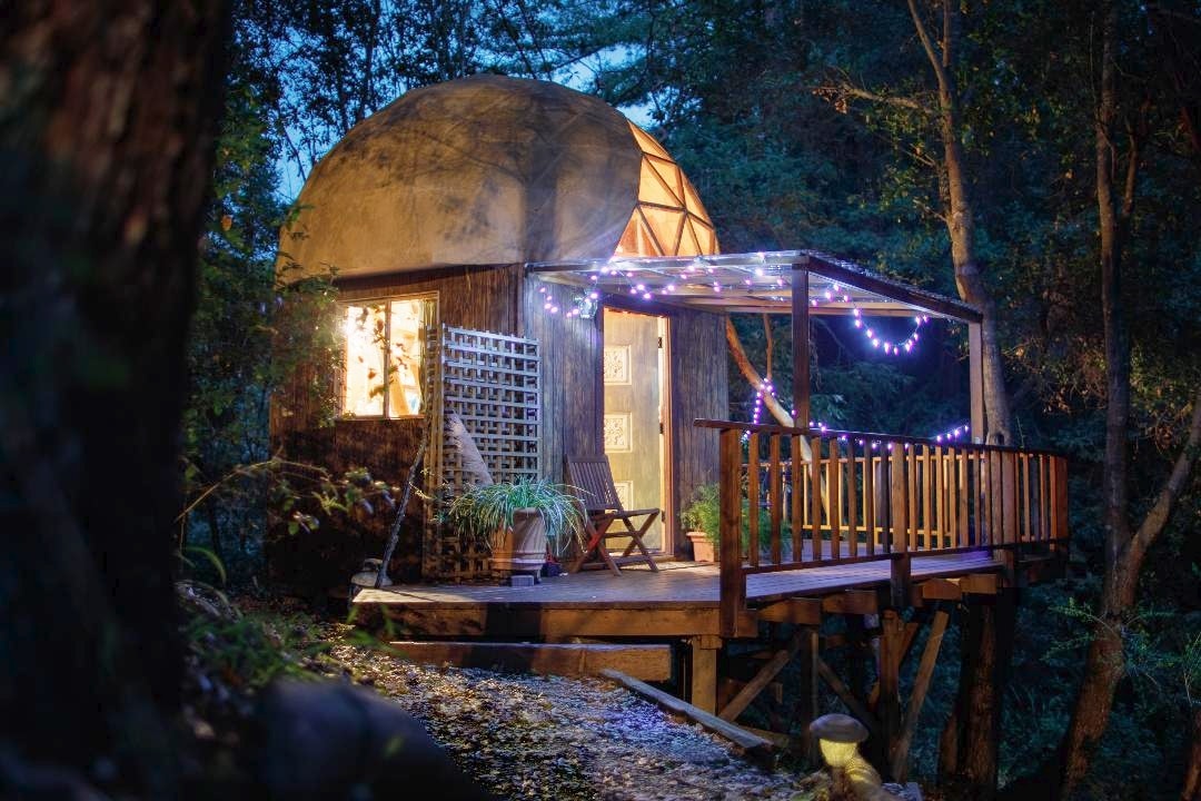 A geodesic dome cabin is perched on wooden stilts, surrounded by lush greenery. Soft lights gracefully illuminate the deck, enhancing the inviting entrance. Large windows reveal a warm interior, while a cozy seating area awaits outside for relaxation amid nature.