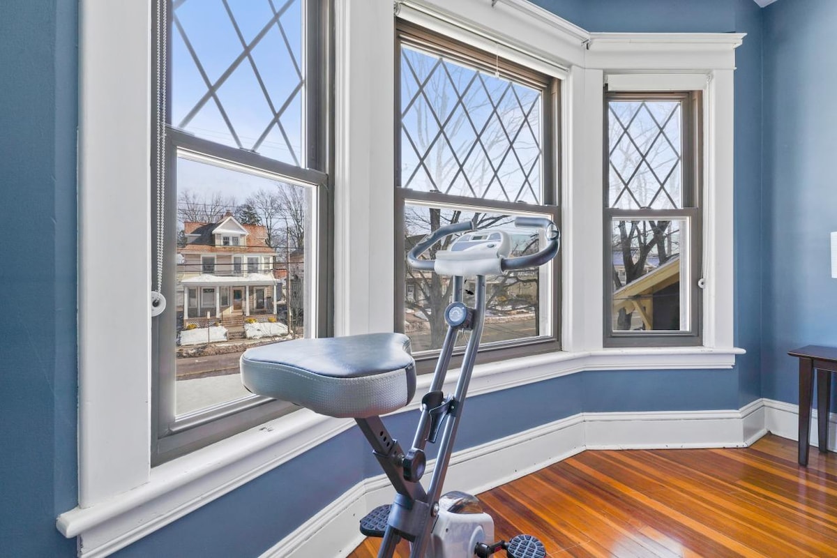 The exercise bike is positioned in front of large bay windows, providing natural light and views of the neighborhood. The blue walls contrast with the warm wooden floor, and the windows feature distinctive diamond patterns, allowing for a bright and open atmosphere.