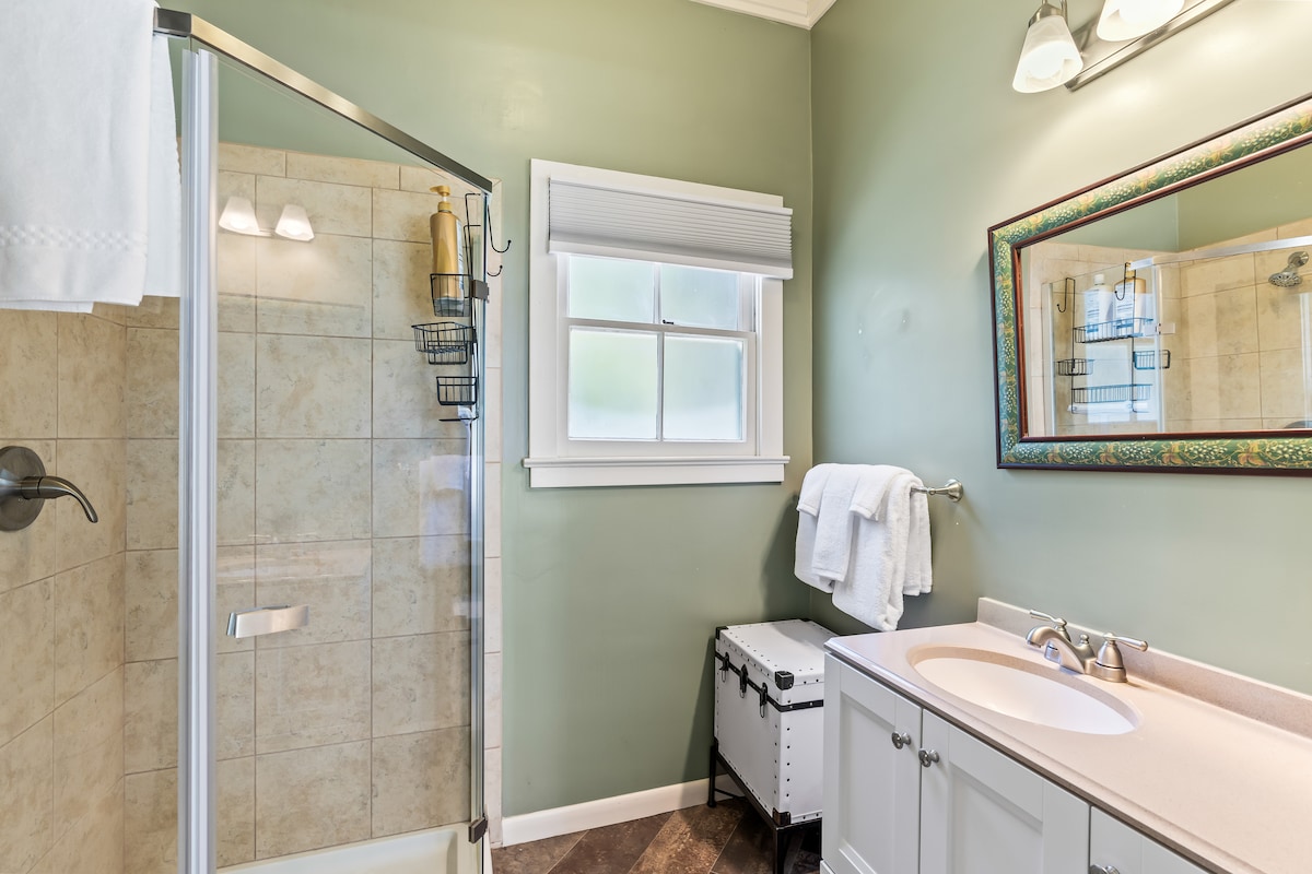 A well-appointed bathroom features a glass-enclosed shower with light tile and a modern sink with storage underneath. A large mirror adds depth, while a window allows natural light to illuminate the space. Towel racks and a stylish storage trunk complete the inviting atmosphere.