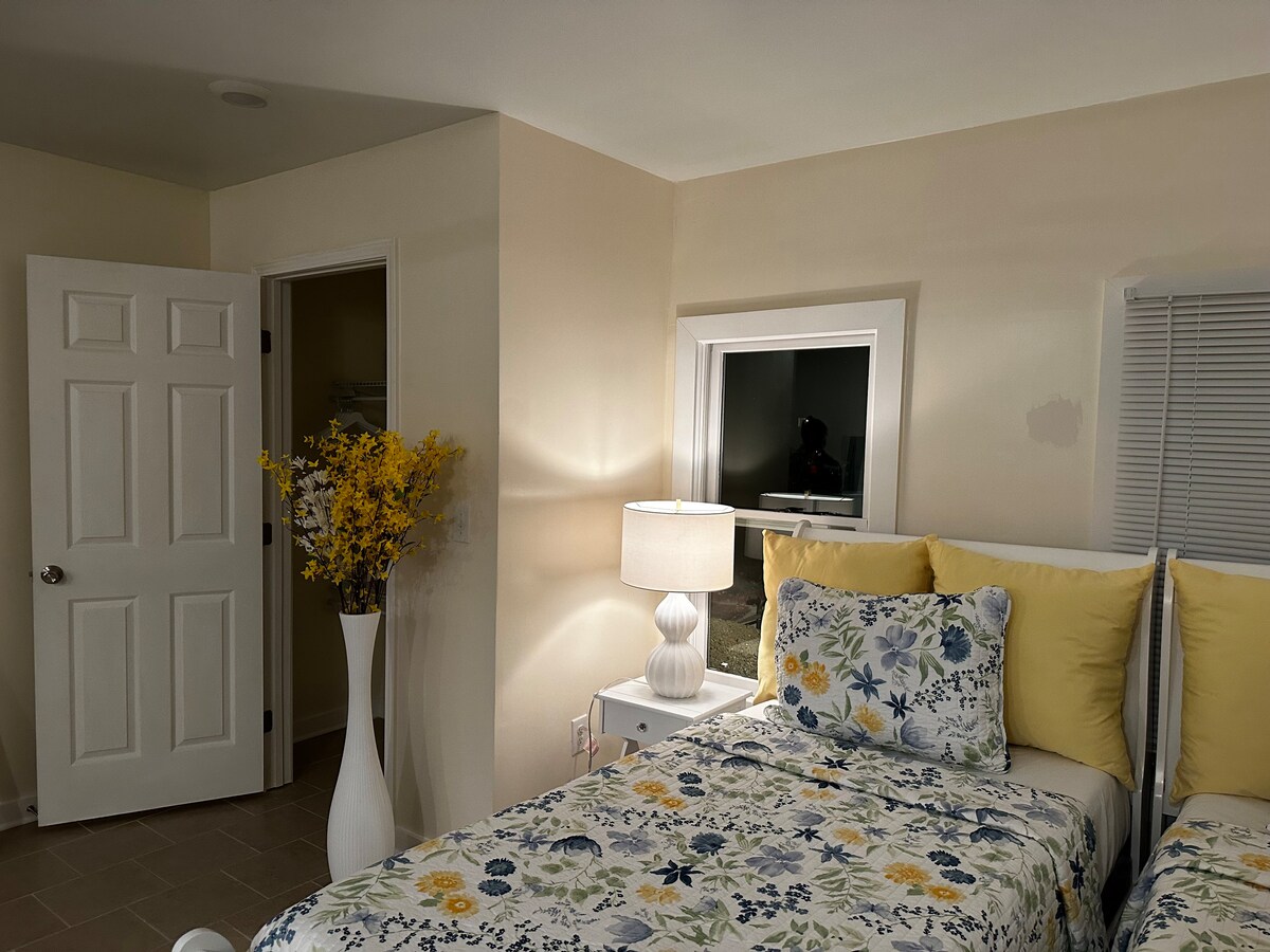 A bright room features two beds dressed in floral-patterned linens. A lamp with a soft glow stands beside one bed. A door leads to a closet, while a window offers a glimpse of the outside. Fresh flowers are displayed in a vase, adding a touch of color.
