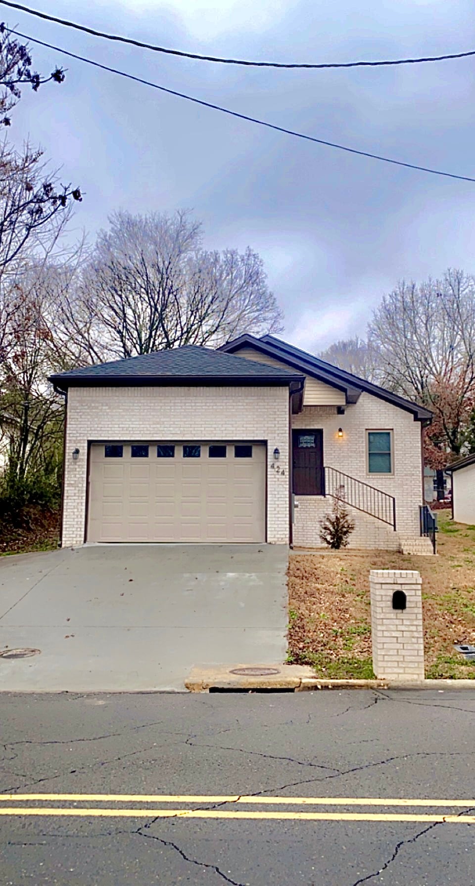 A new construction home is showcased, featuring a light-colored brick exterior with a single-car garage. The sloped driveway leads up to the front entrance, flanked by landscaping and trees. The house has a modern design with a pitched roof and a welcoming front porch.
