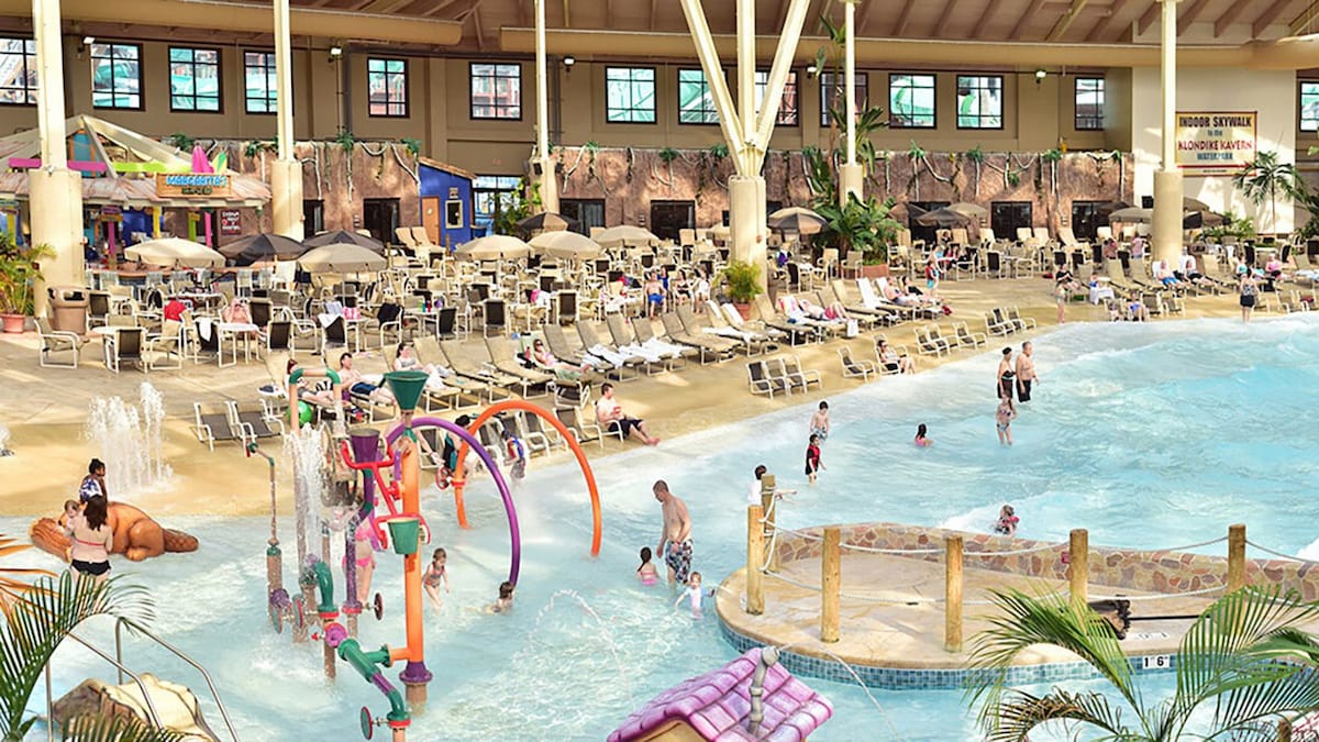 An expansive indoor water park features various water attractions, including playful splash zones and gentle wave pools. Multiple lounge chairs are arranged around the water, providing ample seating for guests. Guests of all ages can be seen enjoying the facilities in a lively atmosphere.