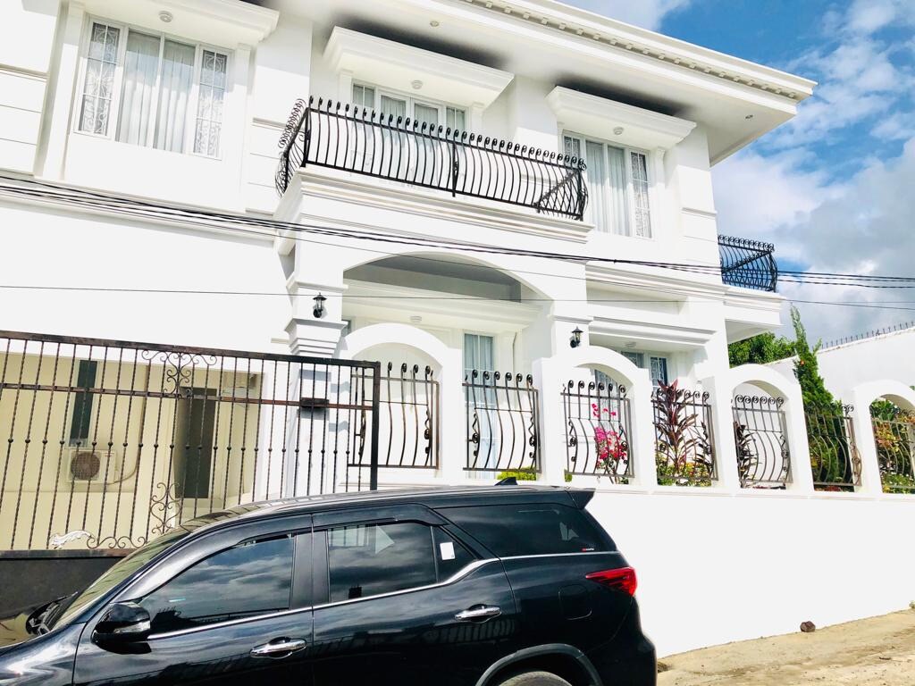A two-story white house is framed by a decorative iron fence, showcasing a modern architectural style. Balconies are visible on the upper level, adorned with black railings. The vehicle parked in front adds to the suburban setting, while lush plants provide a touch of greenery.