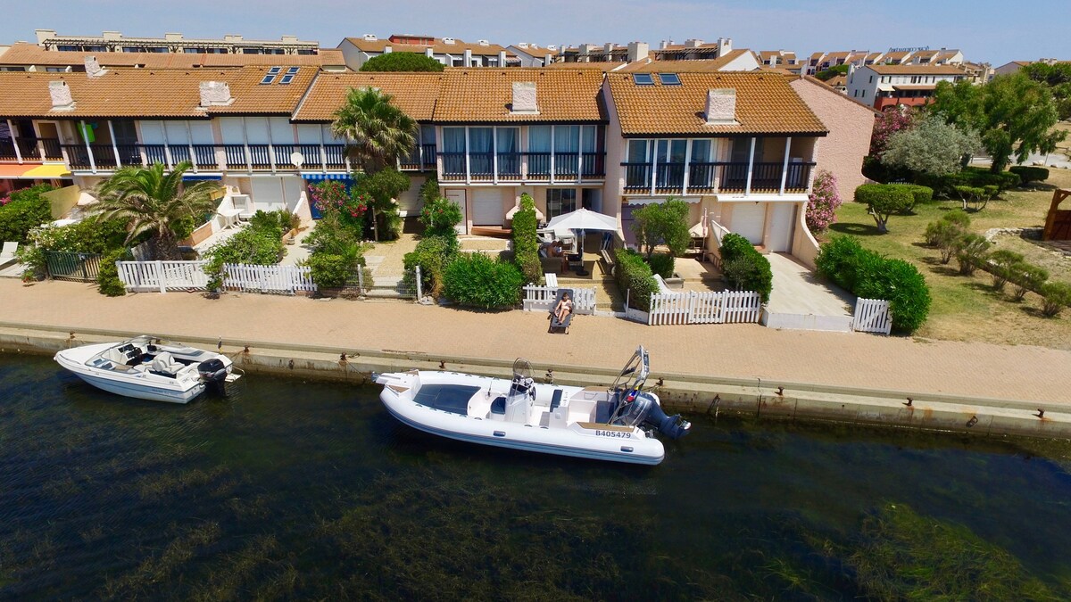 A charming waterside property is visible, featuring a row of homes with large windows and outdoor seating areas. Lush greenery is noted in the gardens, along with boats moored by the water's edge. Sunlight brightens the scene, creating an inviting outdoor space.