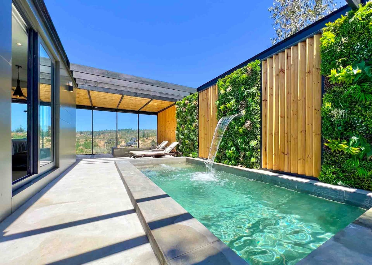 A private pool sits surrounded by lush greenery, with a stylish waterfall feature flowing into the water. Expansive glass walls allow for unobstructed views of the outdoors, while a wooden ceiling provides a warm aesthetic to the serene space.