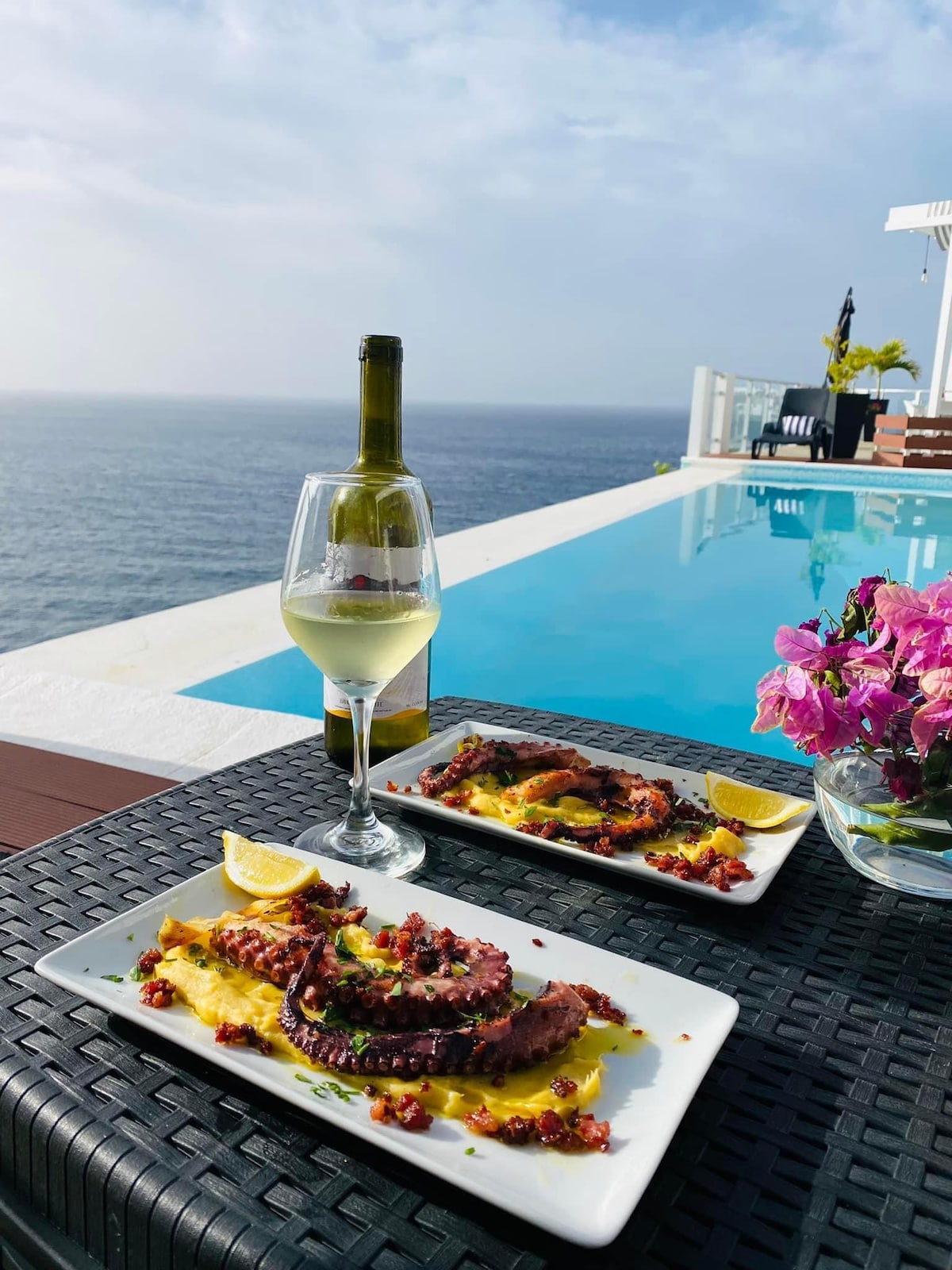A dining setting features two plates of octopus adorned with lemon, accompanied by a glass of white wine. A bottle of wine and a vase with vibrant pink flowers are present on a dark wicker table, with a serene ocean view and swimming pool in the background.