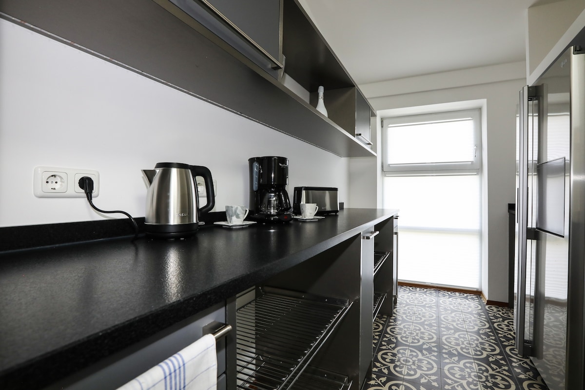 The kitchen features a long, dark countertop with a kettle, coffee maker, and cups neatly arranged. Open shelving above provides storage, while a large window allows natural light to brighten the space. The patterned floor tiles add visual interest.
