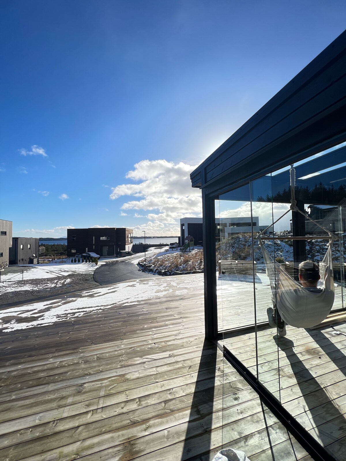 A spacious outdoor deck is depicted, featuring wooden flooring and a hammock. Sunlight filters through large glass doors, highlighting the serene landscape. The sky is clear with scattered clouds, and adjacent buildings are visible in the background, providing a sense of community.